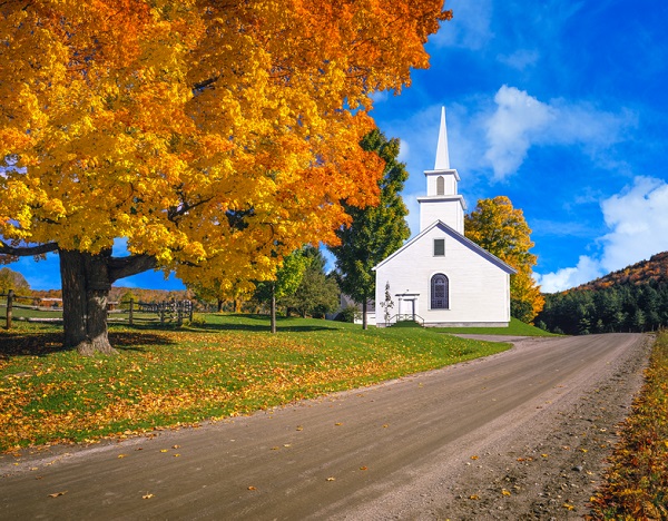 A large sugar maple in full autumn color and a country church fill the foreground leading back to the country side of the Green Mountains, Vermont